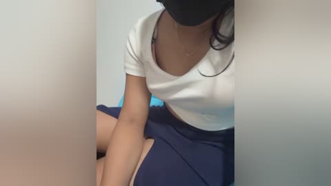 A video of a woman wearing a white crop top, blue skirt, black face mask, seated indoors.