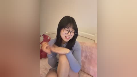 Video of a young East Asian woman with straight black hair and glasses, wearing a blue sweater, sitting on a pink bedspread, holding a red object.