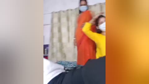A blurry video captures two women in traditional Indian attire, one in a bright orange sari, the other in a yellow blouse and black skirt, interacting in a room with beige curtains and a bed with white sheets.