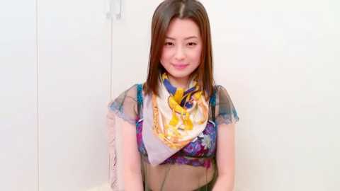 Video of an East Asian woman with straight brown hair, wearing a sheer blue top with a vibrant yellow and purple scarf, against a plain white background.