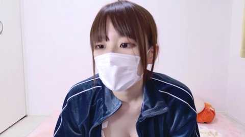 Video of an Asian woman with light skin and brown hair, wearing a white medical mask and a dark blue velvety jacket, sitting on a bed with a pink blanket, in a white-walled room.