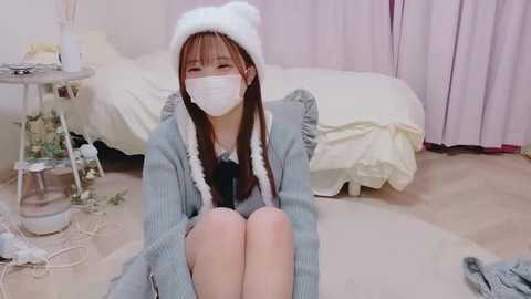 Video of a young Asian woman with long brown hair, wearing a white knitted hat, light blue sweater, white mask, and sitting on a beige carpet in a softly lit, pastel bedroom.