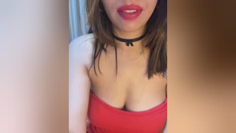 Video of a light-skinned woman with shoulder-length brown hair, wearing a red strapless top that reveals cleavage and a black choker necklace. The background is blurred, focusing on her face and upper body.