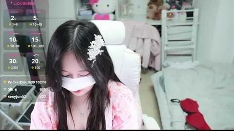 Video of a young East Asian woman with long black hair and a floral headband, wearing a pink robe and a white face mask, sitting on a white chair in a tidy, brightly-lit bedroom with stuffed toys and a bed.
