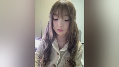 Media: A video of a young Asian woman with long, wavy brown hair, wearing a beige, ribbed cardigan, standing in a softly lit room with a blurred background.