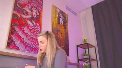 Media: Video of a blonde woman in a grey top, seated in a modern room with vibrant, abstract art on the wall, a black metal shelf, and a dark curtain.