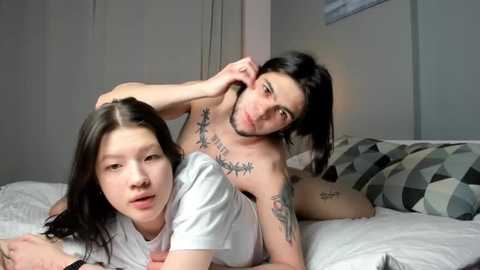 Video of two East Asian women with medium skin tones, one topless, lying on a bed with geometric-patterned pillows.