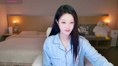 Video of an East Asian woman with long black hair, wearing a light blue striped shirt, sitting on a bed in a modern, beige-themed bedroom with two stuffed animals.