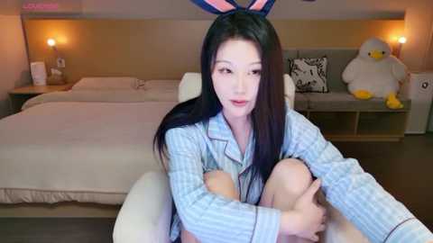 Video of an East Asian woman with long black hair, wearing a light blue plaid pajama top and bunny ears, sitting on a white sofa in a modern, beige-toned bedroom with a bed, two lamps, and a stuffed duck.