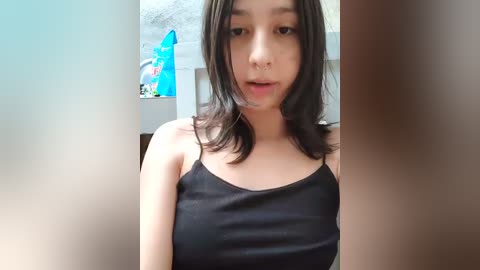 Video of a young Asian woman with shoulder-length black hair, wearing a black spaghetti-strap tank top, standing in a dimly lit room with a blue and white stuffed animal in the background.