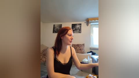 Video of a fair-skinned redhead woman in a black lace camisole, sitting on a bed in a cluttered bedroom with framed photos on the wall and a window with blinds.