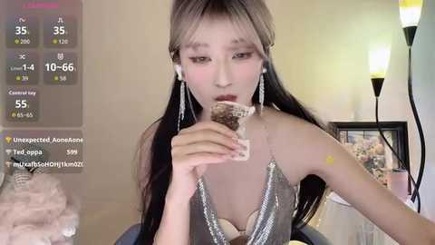 A video of an Asian woman with long black hair, wearing a silver halter top, eating a chocolate bar, in a modern living room with a TV screen showing live stream details.