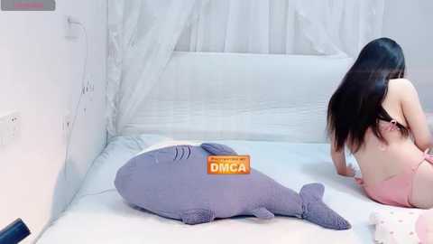 Video of an Asian woman with long black hair, wearing a pink bra, lying on a white bed with a gray dolphin plushie and a white canopy.