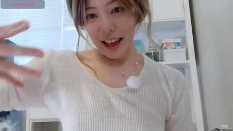 Video of an Asian woman with light skin, wearing a white ribbed sweater, smiling with braces and a septum ring. Background shows a white kitchen cabinet and partially visible blinds.