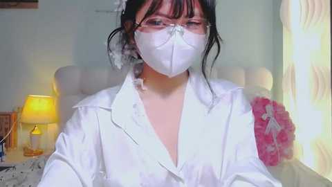 Video of a young Asian woman in a white lab coat with a white mask, standing in a brightly lit bedroom with a yellow lamp and patterned curtains.