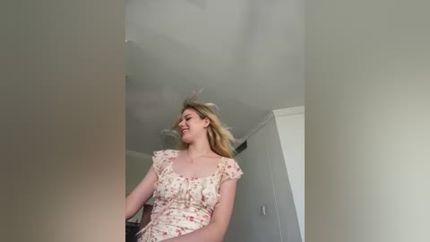 Video of a young, fair-skinned woman with blonde hair, wearing a floral dress, standing in a brightly lit, minimalist room with white walls and a ceiling fan.