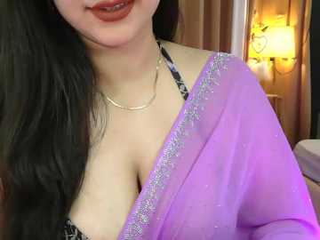 Video of an East Asian woman with long black hair, wearing a purple sari with silver embroidery, revealing a black bra, against a warmly lit, cozy background.