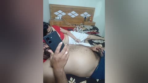Video of a man in a red shirt taking a mirror selfie in a bedroom with a wooden headboard, patterned bedspread, and another person lying on the bed.