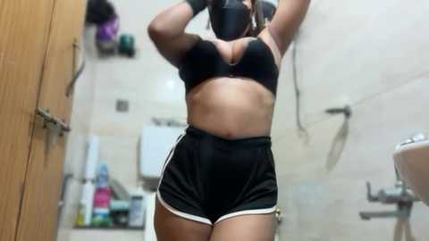 Video of a fit, light-skinned woman with medium-length brown hair, wearing a black sports bra and high-waisted black shorts with white trim, standing in a tiled bathroom with toiletries and a sink in the background.
