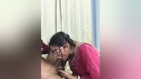 A video shows a South Asian woman with glasses, wearing a pink saree, performing oral sex on a man with a beard. The background features a beige wall and a blue door.