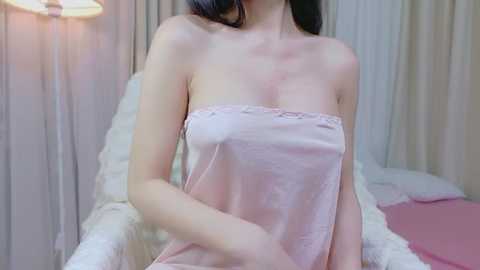 Video of a fair-skinned woman with long black hair, wearing a sheer pink towel covering her breasts, sitting on a bed with white and pink bedding, in a softly lit, cozy bedroom.