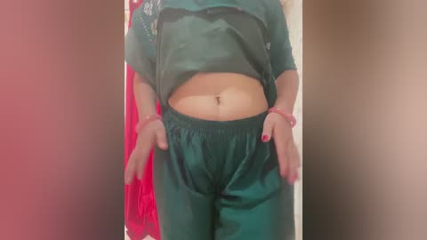 Video of a person with a medium skin tone, wearing a teal satin pyjama set with a red sash, standing in a dimly lit room with a blurred background.