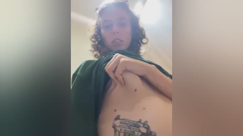 Video of a light-skinned woman with curly hair, lifting a green shirt to reveal a tattoo of a tree on her lower abdomen. Background is blurred, with soft lighting.