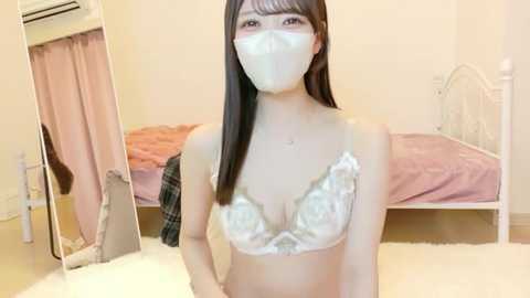 Video of a young Asian woman with long black hair, wearing a white lace bra, face mask, and a plaid shirt, standing in a pastel-toned bedroom with a bed and mirror.