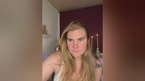 Video of a blonde woman with light skin, wearing a white tank top, sitting in a dimly lit room with purple walls. She has tattoos on her shoulder and arm, and there are candles on a table behind her.