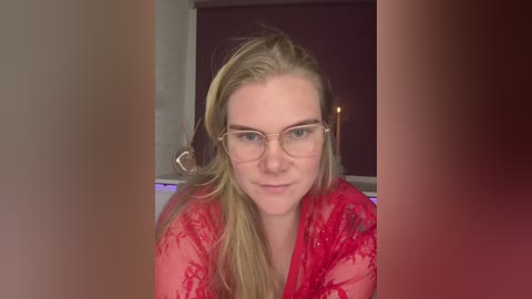 A video of a fair-skinned woman with long, blonde hair, wearing wire-framed glasses and a red lace robe, sitting indoors.