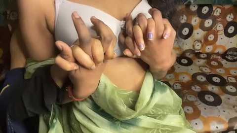 A video of a woman with medium brown skin, wearing a green blouse and white bra, playfully pinching her own nipple. She has a dark complexion and short hair. Background shows a patterned blanket.