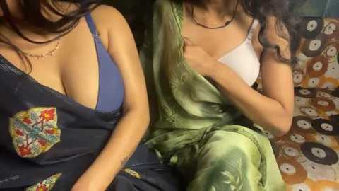 Video of two women, one in a blue tank top, the other in a green saree, sitting closely, with visible cleavage and a patterned couch background.