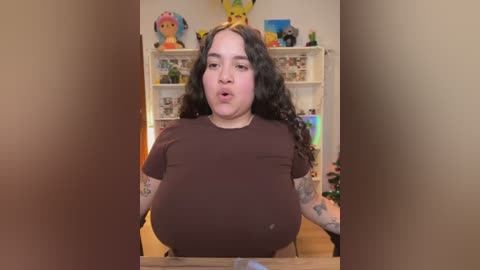 Video of a plus-sized woman with long curly brown hair, wearing a brown shirt, in a colorful, cluttered room with toy figures and shelves in the background.