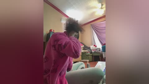 Video of a person with a voluminous afro in a pink robe, standing in a cluttered bedroom with a green bed and pink curtains.