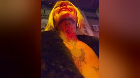 Video of a blonde woman with vivid red face paint, lying back, exposing a tattoo on her neck and chest. Background is blurred, giving a dramatic, intense effect.