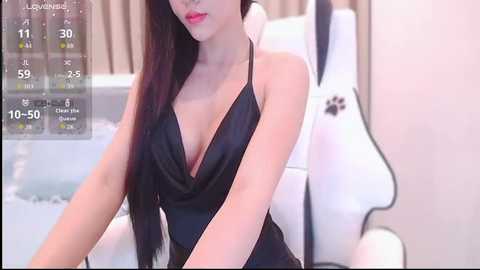 A video of a young Asian woman with long, straight black hair and fair skin, wearing a black halter-neck top, seated in a modern, white bathroom with a digital display showing time and temperature.