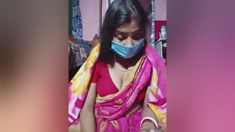 Video of a South Asian woman with medium skin tone and black hair, wearing a pink saree, blue mask, and white bracelet, seated in a modest indoor setting.