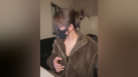Video of a woman in a brown fuzzy jacket, black mask, and glasses, adjusting her hair, standing indoors with beige walls and a window with vertical blinds.