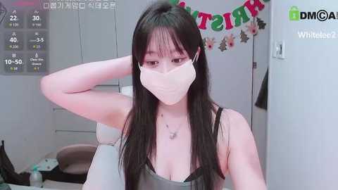 A video of an Asian woman with long black hair and a pale complexion, wearing a mask, sitting in a room with a \"MOMA\" banner and a calendar.