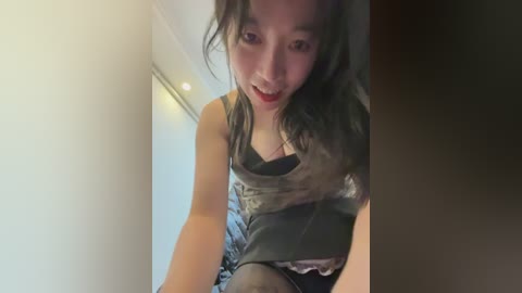 Video of a young Asian woman with long black hair, wearing a revealing black leather top, sitting on a bed, smiling.