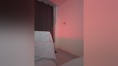 A video of a dimly lit room with white bedding and a dark, curtained window. The walls are painted light beige, and the room is bathed in soft red and pink hues from a hidden light source.