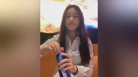Video of a young Asian woman with long dark hair, wearing glasses, a white button-up shirt, and a blue drink bottle, sitting indoors with a colorful abstract painting behind her.