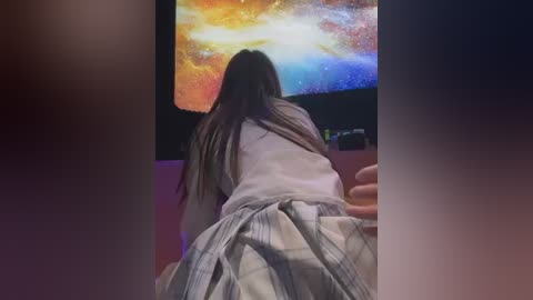 Video of a person with long, dark hair, wearing a white shirt and plaid skirt, kneeling on a couch, facing a colorful, abstract starry sky TV screen.