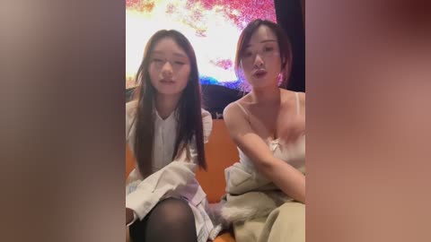 Video of two young East Asian women with fair skin, one in a white dress, the other in a beige outfit, seated on a couch in front of a colorful abstract painting.
