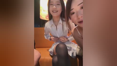 Video of two young Asian women smiling, one wearing a white blouse and black tights, sitting on a brown couch, with a vibrant painting in the background.