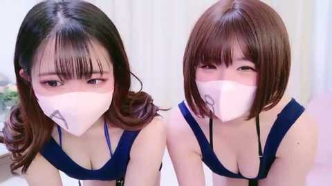 Video of two Asian women with short bob haircuts and white masks, wearing blue tops, looking down. Background shows a white room with a green plant.
