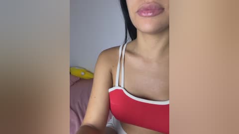 Video of a young woman with long black hair, wearing a red bra with white trim, partially visible. She has light skin, and her lips are slightly parted. Background features a pink bedspread and a yellow stuffed toy.