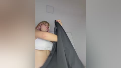 A video of a blonde woman in a white crop top, holding a large, dark gray blanket, in a minimalistic, white-walled room with a small vent.