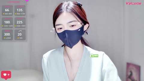 Video of an Asian woman with long dark hair, wearing a dark blue medical mask and white lab coat, standing in a sterile, white-lit medical room.