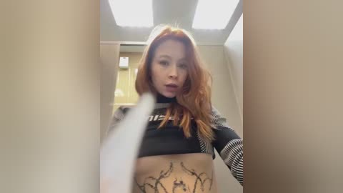 Video of a young woman with long red hair, wearing a black crop top, revealing a tattoo on her midriff, standing in a hallway with beige walls and fluorescent lighting.